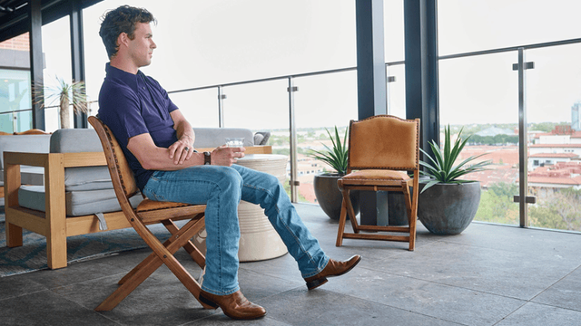 A man sitting in a chair on a rooftop bar, wearing the Amado in brown.