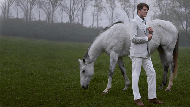 A man wearing a grey suit standing next to a grey horse in a pasture, wearing the Lenado 12” in brown.