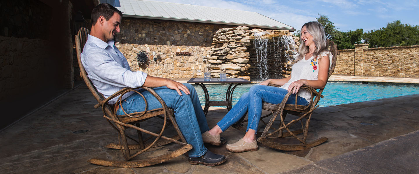 Man and women sit in front of pool in casual Tony Lama slip-ons in tan and blue.