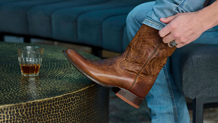 Man wearing a pair of Derby western boots in brown with a glass of whiskey on a coffee table.
