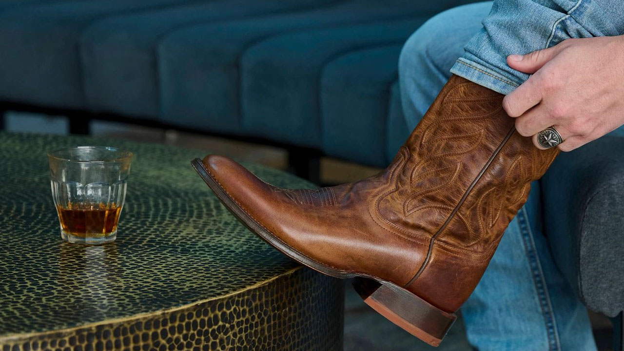 Man wearing a pair of Derby western boots in brown with a glass of whiskey on a coffee table.